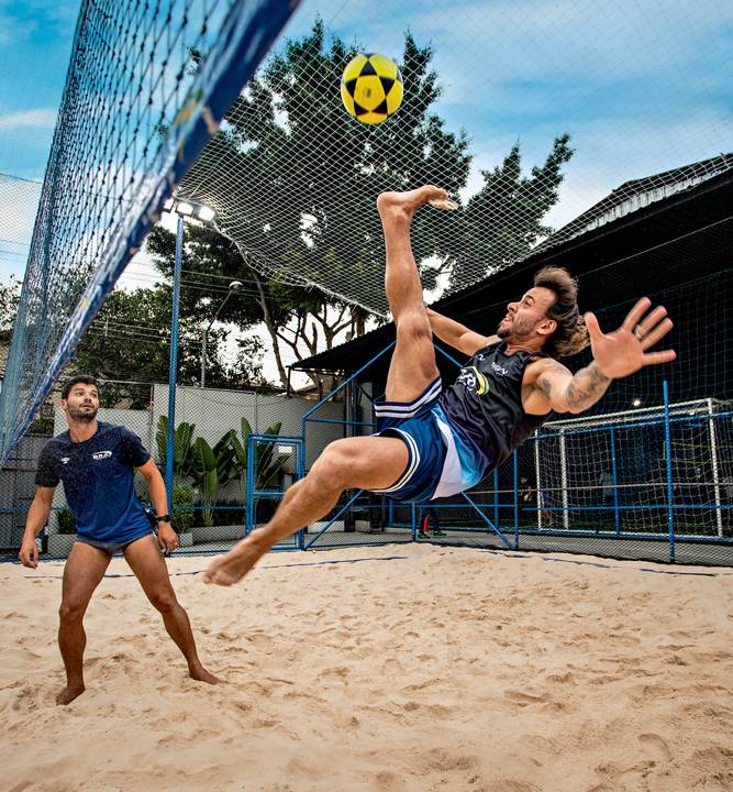 A imagem mostra um jogador de futevolei, em uma quadra de areia, pulando para trás e elevando sua perna direita para acertar a bola no alto.