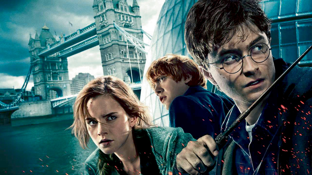 A imagem mostra os três protagonistas do filme Harry Potter com um castelo ao fundo, da ambientação do filme. Eles estão com uma expressão de seriedade.