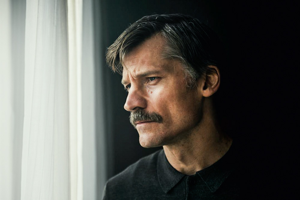 A imagem mostra Nikolaj Coster-Waldau, só de bigode, olhando preocupado pela janela