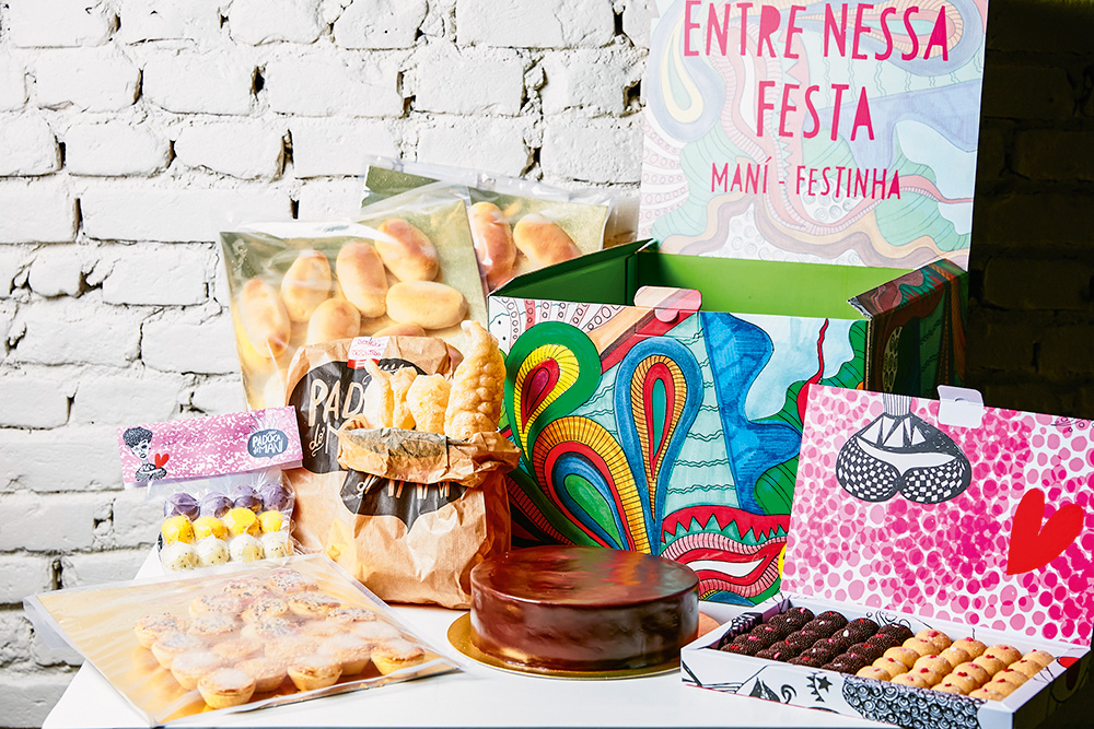 Foto do kit "Maní-Festinha" da Padoca do Maní sobre mesa, com bolo de chocolate à frente, caixa de brigadeiros à direita, pães e salgadinhos à esquerda.