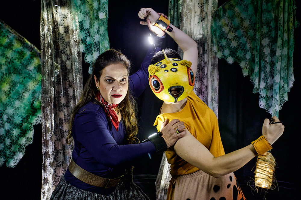 Foto mostra um espetáculo em palco. Uma mulher, vestida de caçadora está ao lado de uma atriz fantasiada de onça pintada. No fundo, cenário de floresta