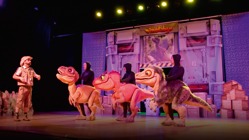 Em um palco de teatro, um homem vestido de paleontólogo conversa com três atores fantasiados de dinossauros filhotes