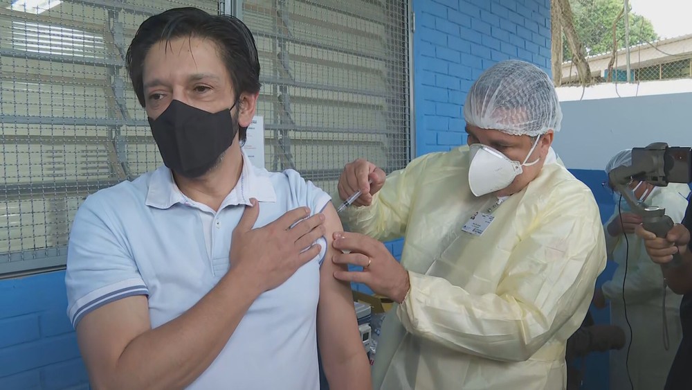 ricardo nunes com uma das mãos no peito enquanto recebe a vacina no outro braço, ministrada por uma enfermeira