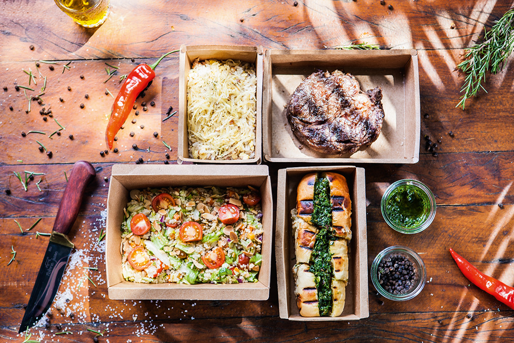 Foto aérea de embalagens de delivery do Pobre Juan, com arroz biro-biro, carne grelhada, choripán e salada.