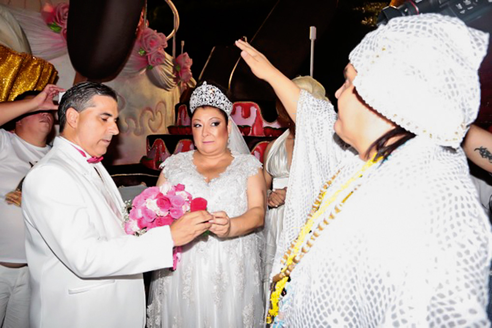 nosso-louco-amor-casamento-carnaval-rosas-de-ouro-angelina-cerimônia-pai-de-santo sérgio e angelina de mãos dadas enquanto pai de santo realiza cerimônia com o braço levantado