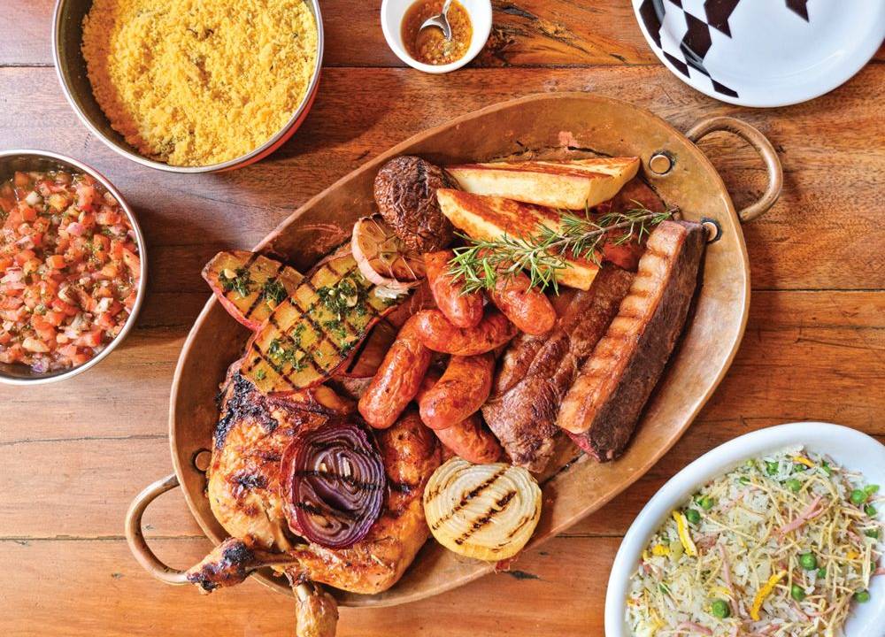 Panela de cobre com múltiplas variedades de carne sobre mesa de madeira ao lado de porções de arroz biro-biro, farofa e vinagrete.
