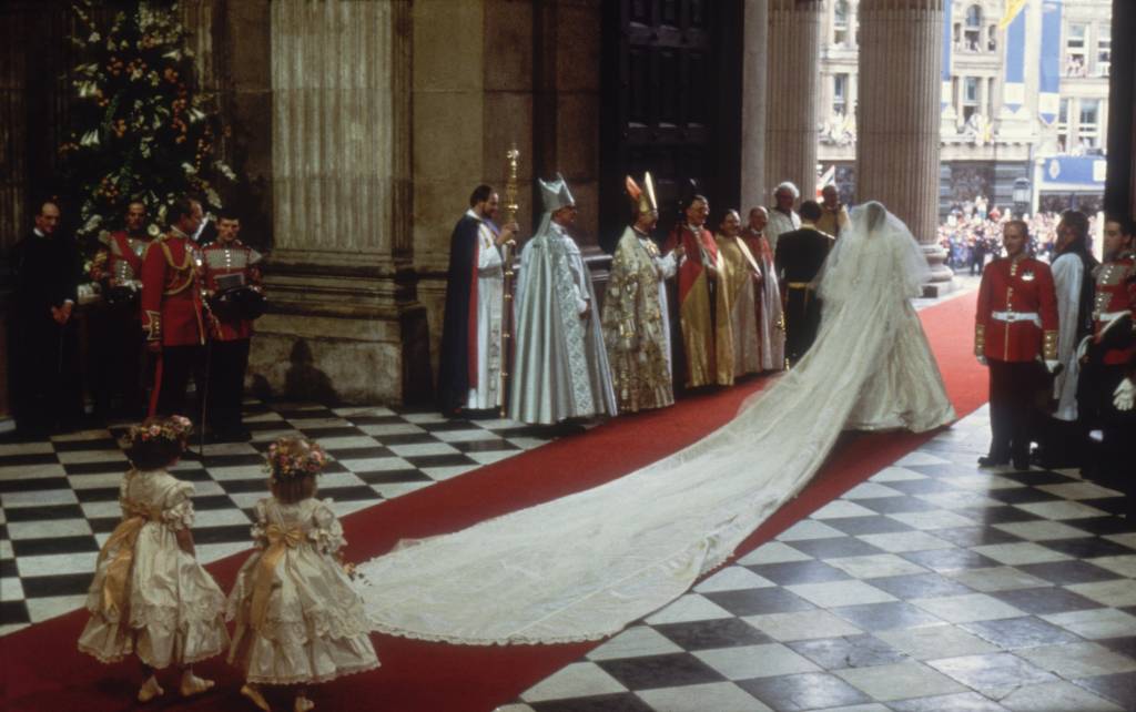 lady di e principe charles entram na igreja sobre um tapete vermelho para a cerimônia. pessoas clericais estão ao redor na porta e as damas de honra carregam o final de seu véu