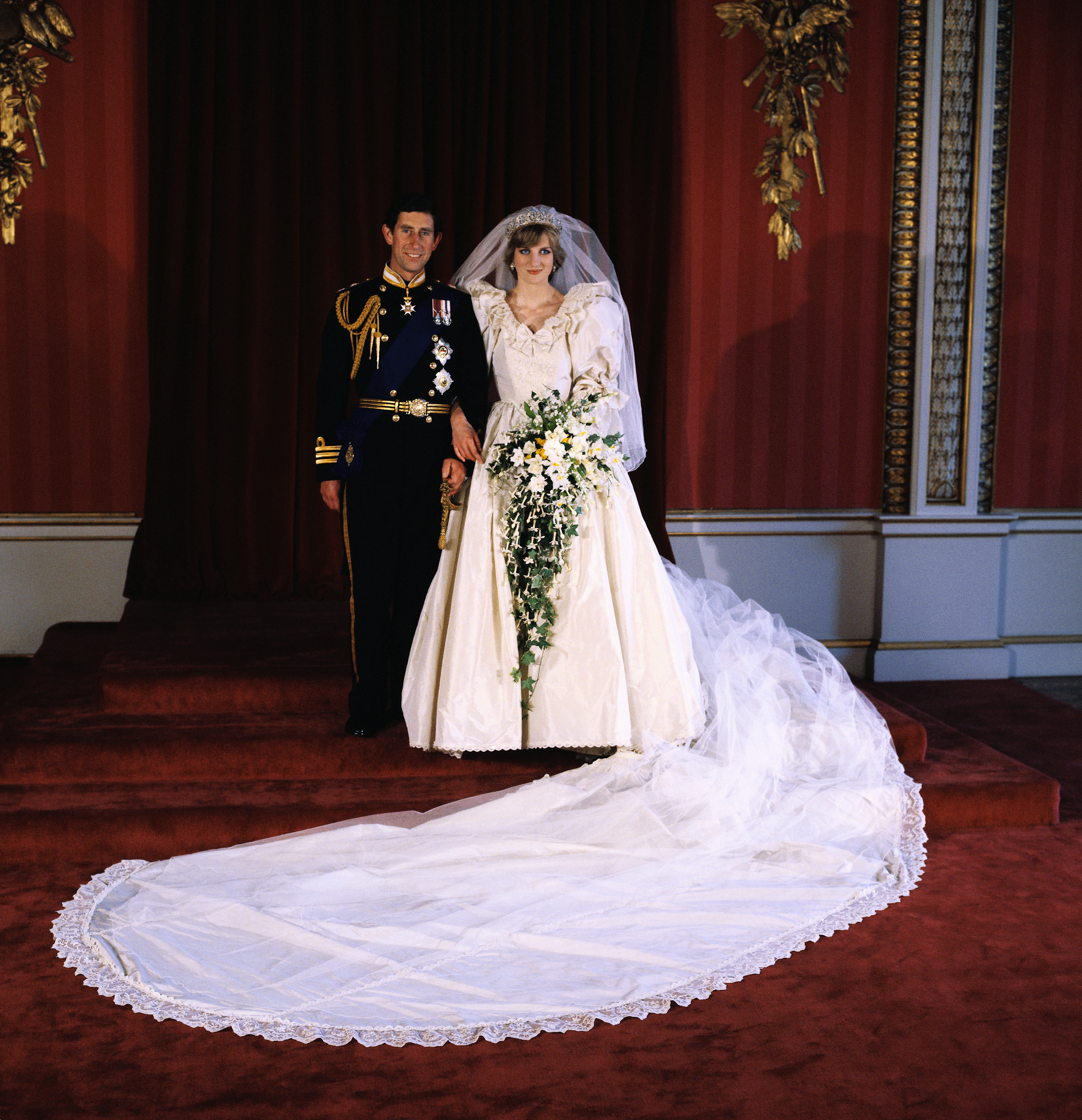 Príncipe Charles e Princesa Diana posando para a foto oficial do casamento, feita pelo Lorde Lichfield no Buckingham Palace, após cerimônia na Catedral de São Paulo, em Londres. eles estão com as vestes do casamento em uma sala com tons majoritariamente vermelhos