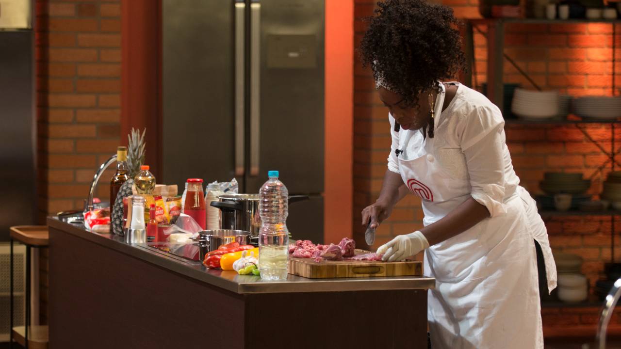 Participante do MasterChef Brasikl Cecília Ramos cortando ingredientes sobre tábua de madeira em bancada do programa.