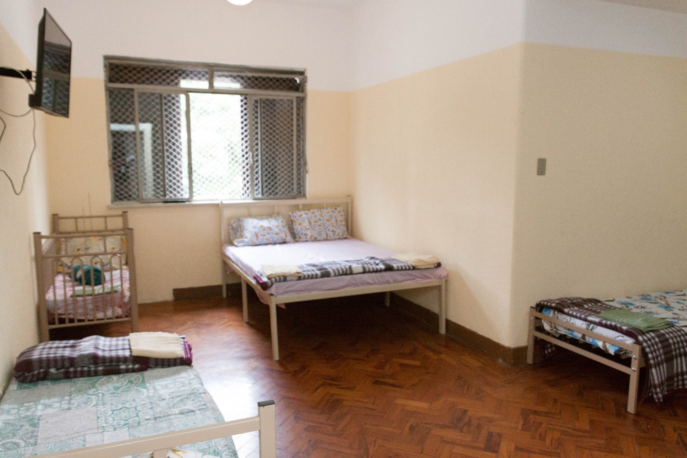 A imagem mostra um quarto no abrigo da prefeitura. É possível ver uma cama de casal, um janela, um berço e outra cama um pouco menor