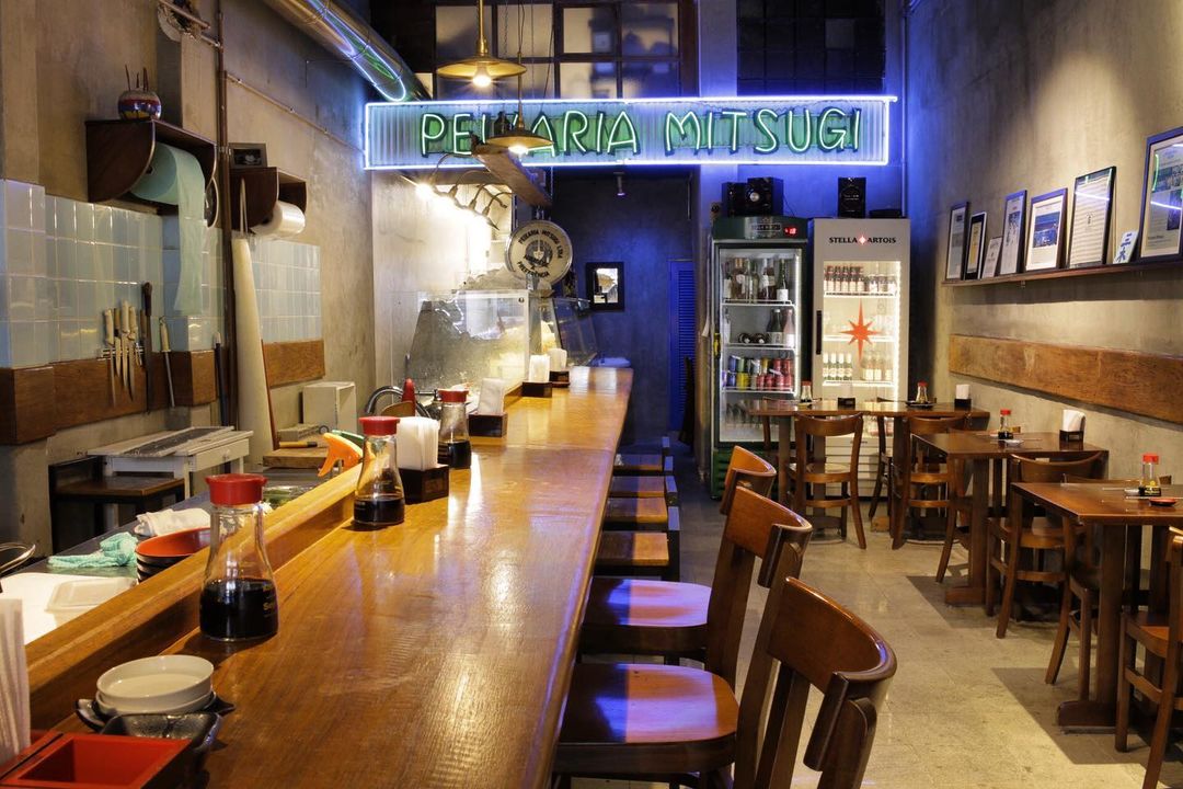 Salão da Peixaria Mitsugi, com balcão e banquetas á esquerda, mesinhas à direita e geladeiras de bebida ao fundo sob letreiro luminoso com o nome da peixaria.