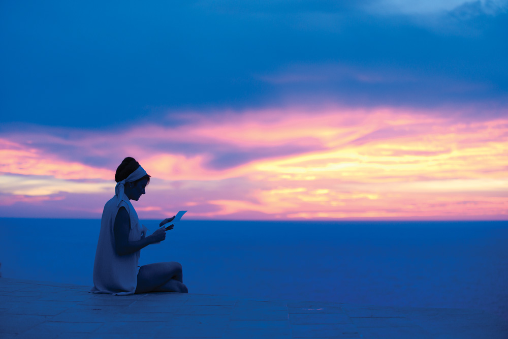 A imagem mostra uma mulher lendo uma carta, durante o amanhecer, em uma imagem bem escura. Ela está sentado em um chão de concreto mas é possível ver o mar ao fundo.