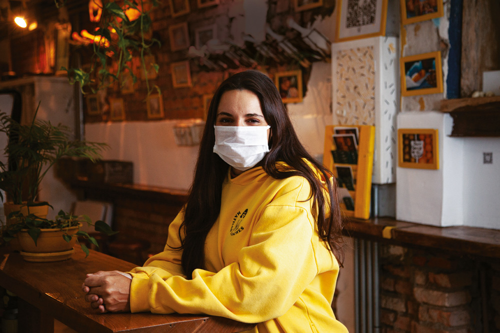 A chef Renata Vanzetto sentada, de moletom amarelo e máscara, com os braços apoiados sobre messa de madeira.