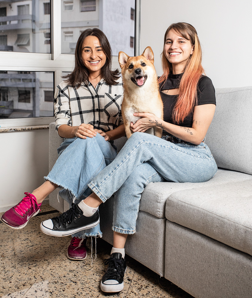 A imagem mostra as duas amigas sentadas em um sofá, com o cachorro no colo, sorrindo para a câmera.