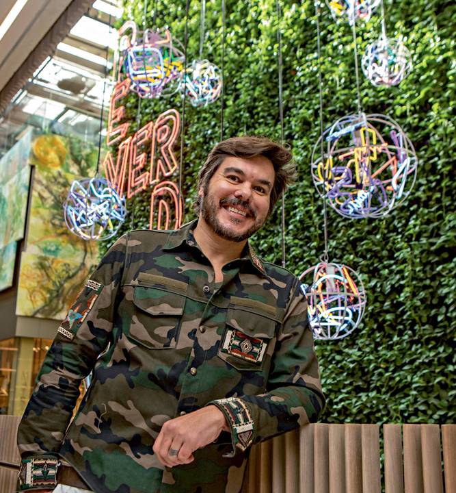 Alê Jordão, artista especializado em peças feitas com luzes neon, posa apoiado em muro de corredor do CJ Shops. Veste camisa com estampa militar e sorri. Ao fundo, esferas compostas por tiras de luzes em neon aparecem iluminadas em frente a parede coberta por plantas.
