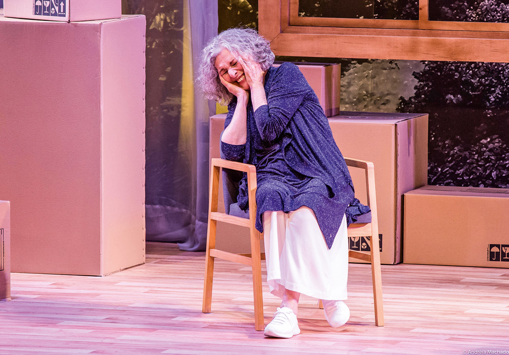 Mulher com expressão de tristeza com as mãos no rosto está sentada em uma cadeira no palco de um teatro