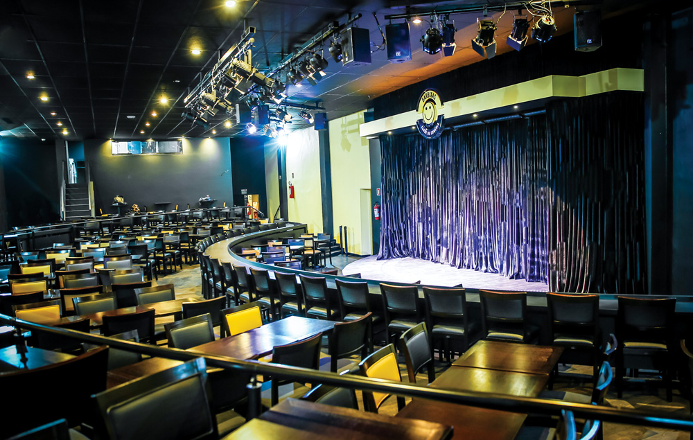 Foto do espaço do Clube Barbixas de Comédia, com um palco e mesas e cadeiras vazias na platéia