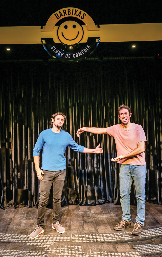 Dois homens fazem pose de abraço, mas com distanciamento social, em um palco de um clube de comédia. Eles estão sorrindo