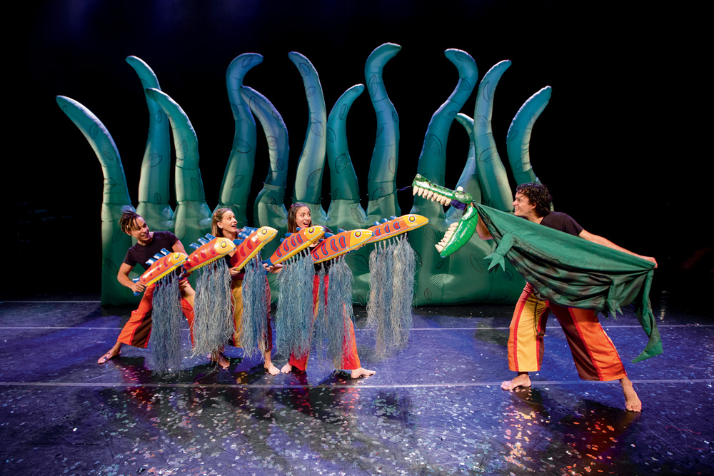 Três atores manuseiam bonecos de peixes e outro de um jacaré em um palco de teatro