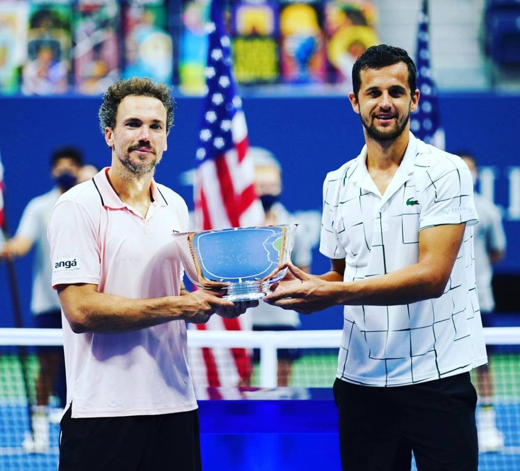 A imagem mostra Bruno Soares e Mate Pavic segurando o troféu do US Open em uma quadra de tênis.