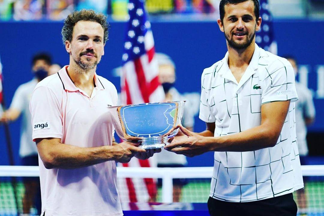 A imagem mostra Bruno Soares e Mate Pavic segurando o troféu do US Open em uma quadra de tênis.