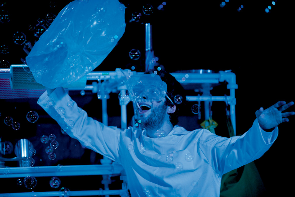 Homem em uma peça tem instrumento de proteção na cara enquanto olha animado para bolinhas de sabão. Imagem tem forte luz azul