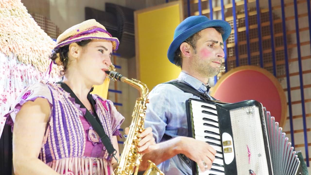 Uma mulher toca um saxofone, vestida de rosa, ao lado de hum homem que toca sanfona, vestido de azul