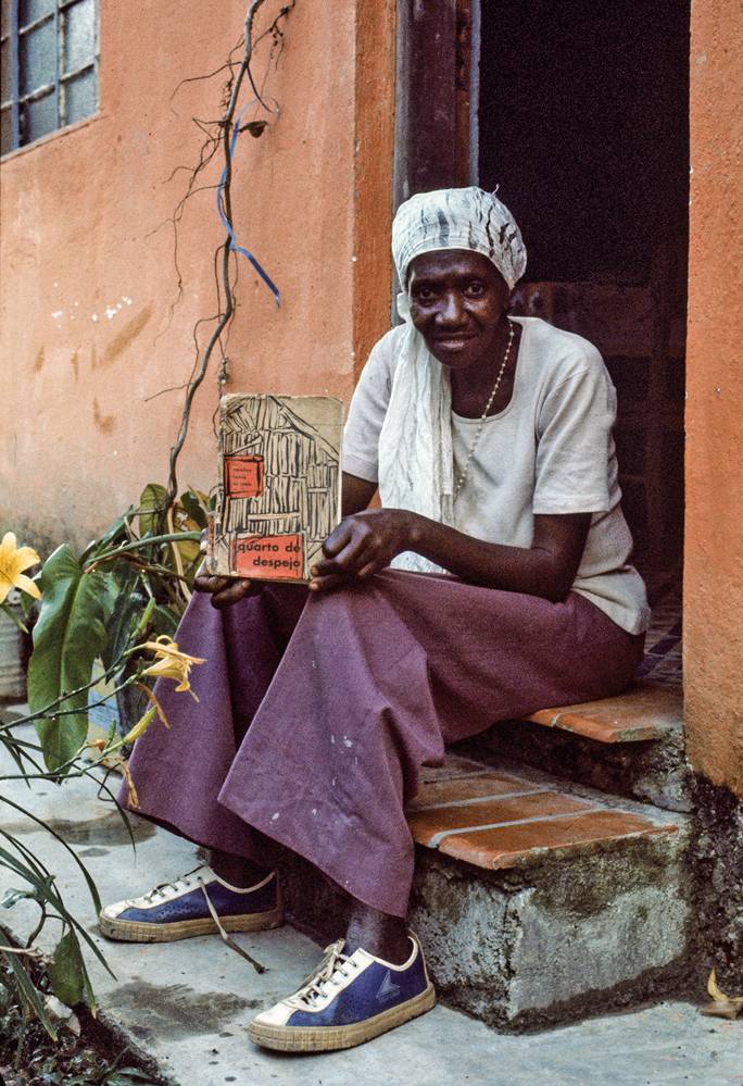 Carolina Maria de Jesus sentada na porta de uma casa, com seu livro 