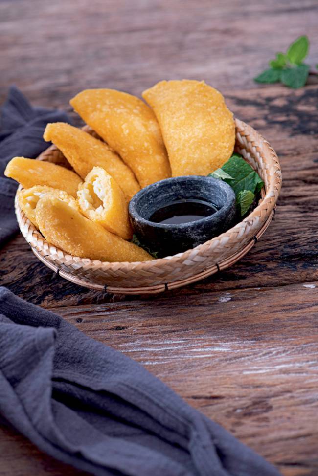 Cestinha de palha com mini pastéis amarelados junto de potinho com melaço sobre mesa de madeira.