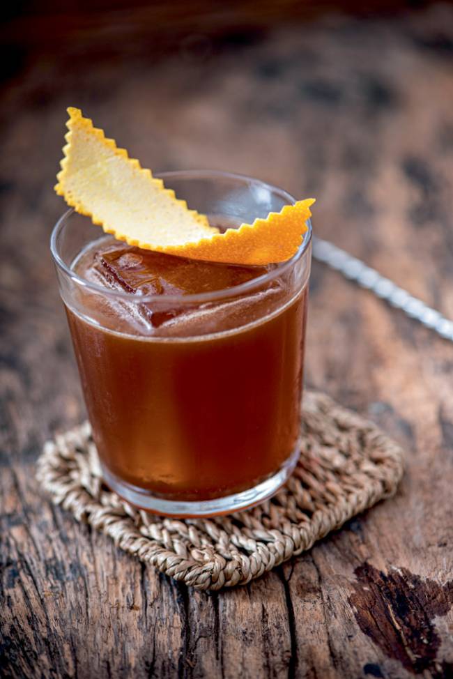 Drinque de cor caramelo com gelão quadrado coberto por casca de laranja sobre porta-copos em mesa de madeira.