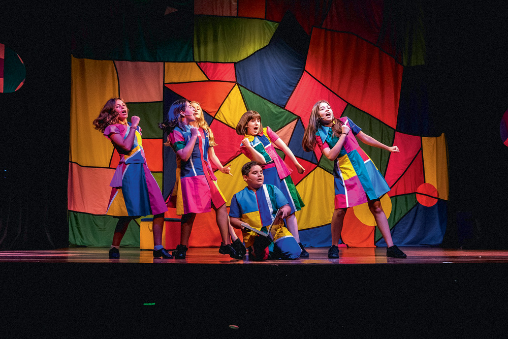 Seis crianças em cena, vestidas com a mesma roupa, de formas e coloridas, dançam em um palco de teatro