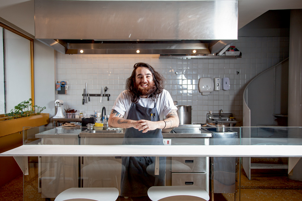 O chef, Raphael Vieira, usando uma camiseta branca e um avental de cor cinza por cima, posa na cozinha de seu restaurante.
