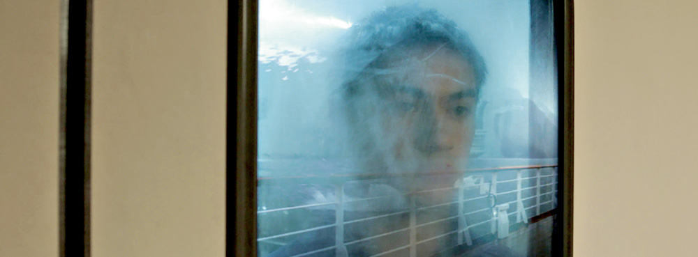 A imagem mostra foto de uma janela, muito embaçada, em que é possível ver parcialmente o rosto do protagonista do longa com uma expressão neutra.