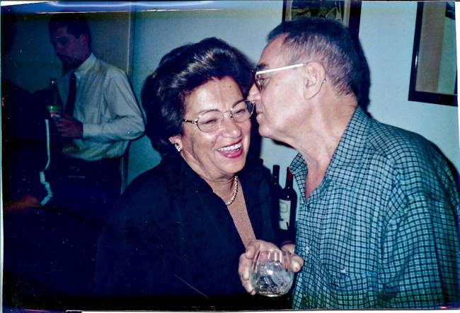 Creusa e Decio – Hamburguinho Foto azulada de Creusa e Décio Silva, fundadores do Hamburguinho, em festa, com ele à direita dando um beijo na bochecha dela.