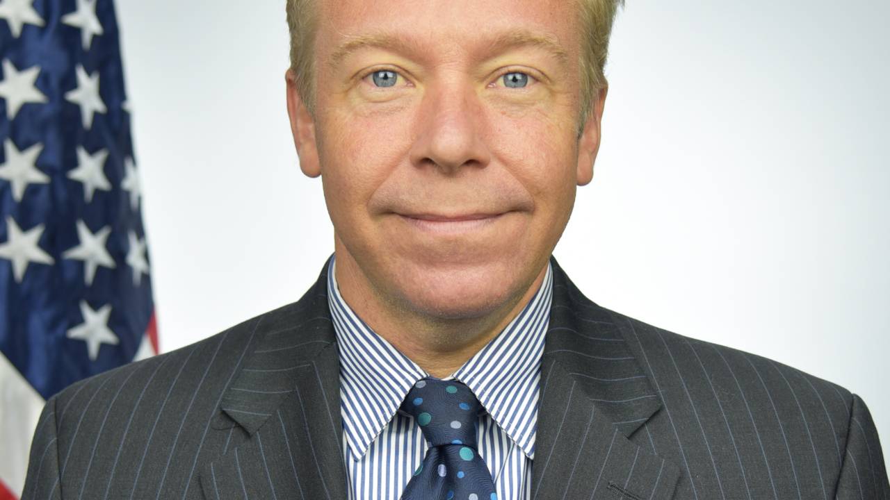 David Hodge em foto 3x4 sorri vestindo terno, gravata azul escuro e cabelos loiros. Ao fundo, a bandeira americana.
