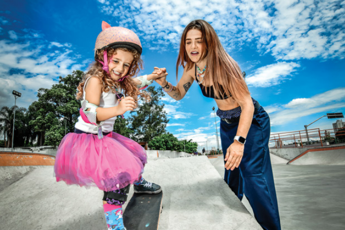 Uma menina equipada com proteção andando de skate de mãos dadas com uma mulher