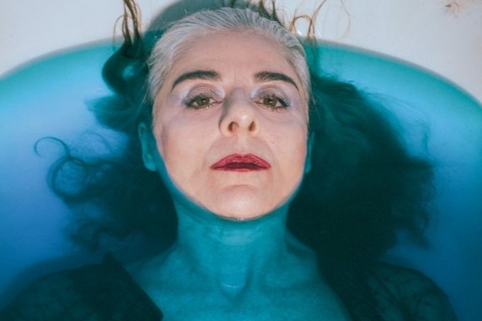 Mulher grisalha está deitada em uma banheira com água azul, só a cabeça dela está para fora