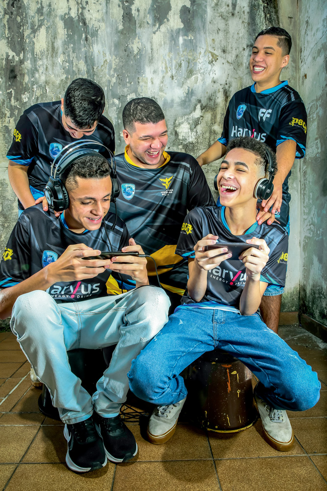 A imagem mostra Cauê, Felipe, Nycolas, Davi e Ryan Barbosa, todos rindo enquanto jogando Free Fire no celular. Todos estão com o uniforme preto da equipe.