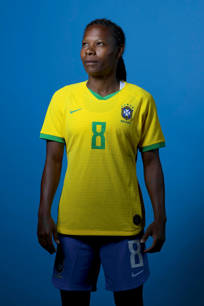A imagem mostra Formiga, com uniforme da seleção brasileira, em um fundo azul. Ela olha para cima.