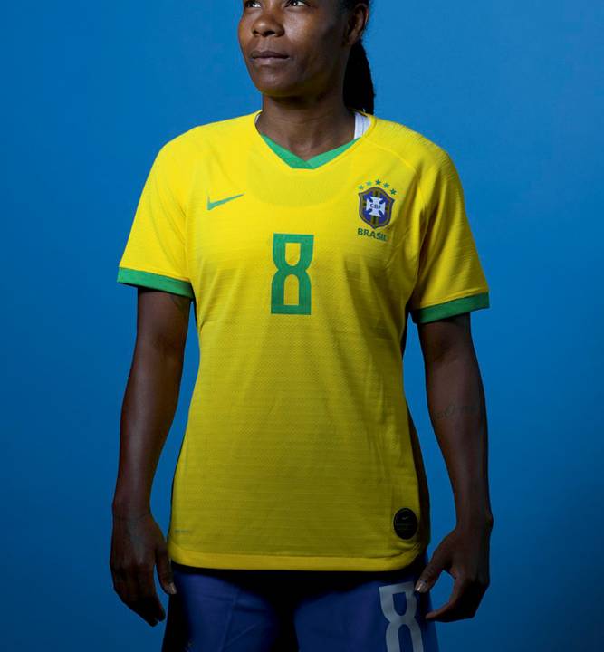 A imagem mostra Formiga, com uniforme da seleção brasileira, em um fundo azul. Ela olha para cima.
