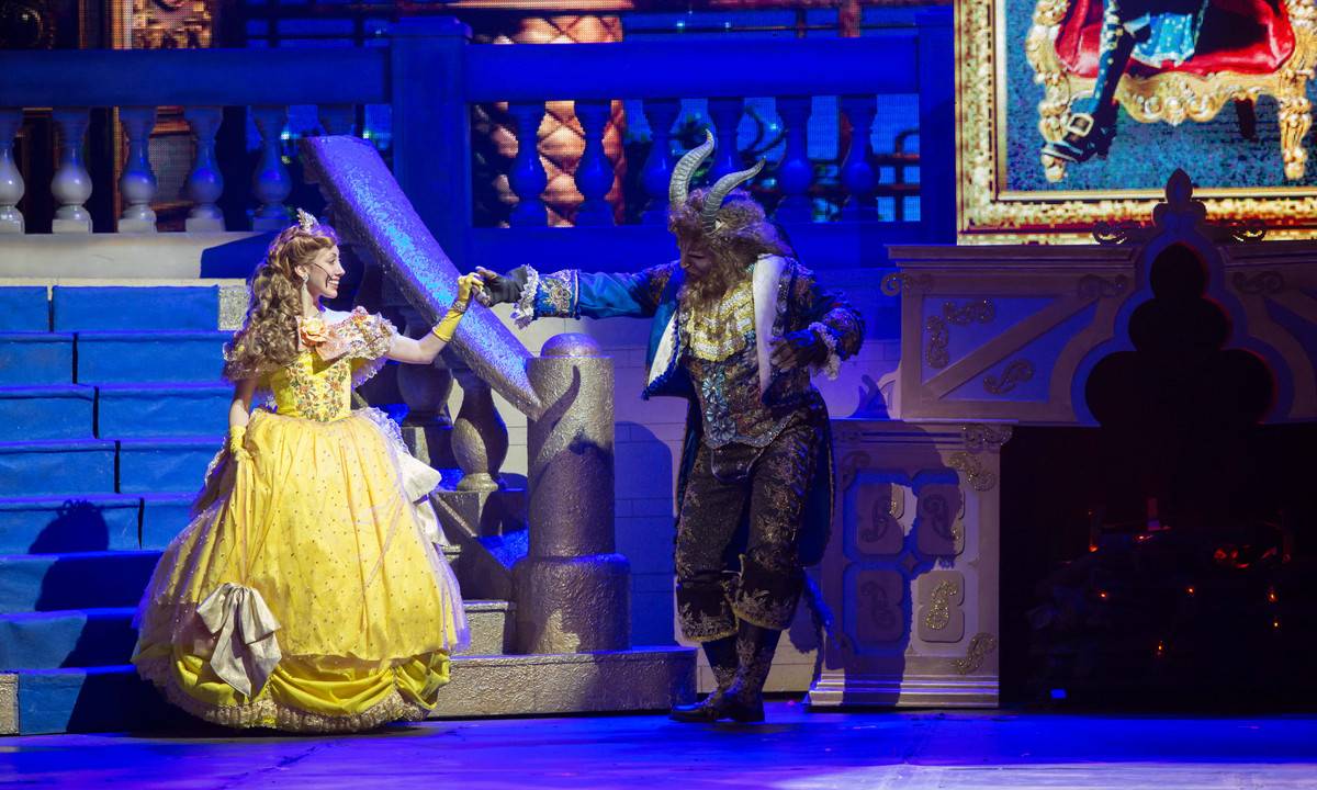 Em um palco de teatro, uma atriz vestida de amarelo como a princesa Bela, dança com um ator vestido de Fera. Luzes azuis iluminam o palco