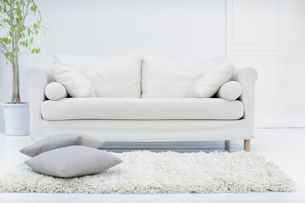 Uma sala de estar com tons de branco. Tem um sofá, duas almofadas no chão e um tapete de pelinhos