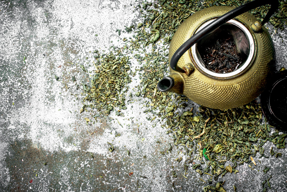 Chá verde em um bule sobre um fundo rústico. Há folhas do chá espalhadas ao fundo