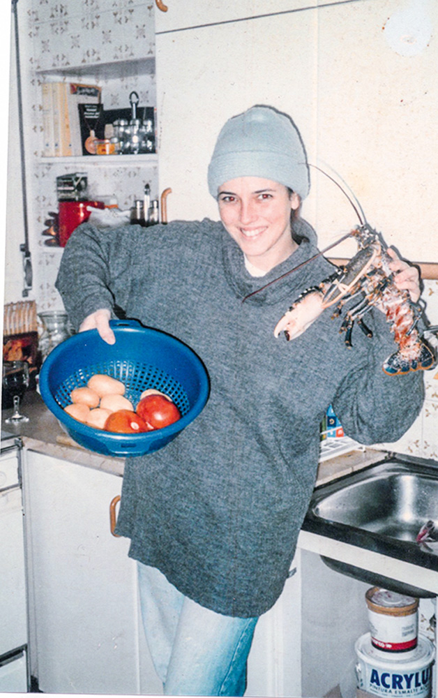 Helena Rizzo de moletom e touca cinzas posando para foto em uma cozinha com uma tigela com tomates e batatas na mão direita e uma lagosta na mão esquerda.