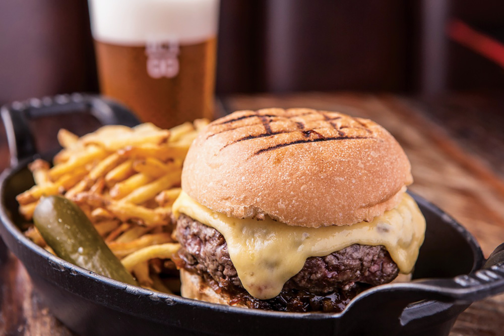 Hambúrguer alto com queijo escorrendo sobre panelinha de metal ao lado de fritas.
