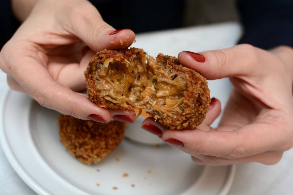 Mãos brancas de unhas pintadas de vermelho partindo croqueta de carne ao meio.