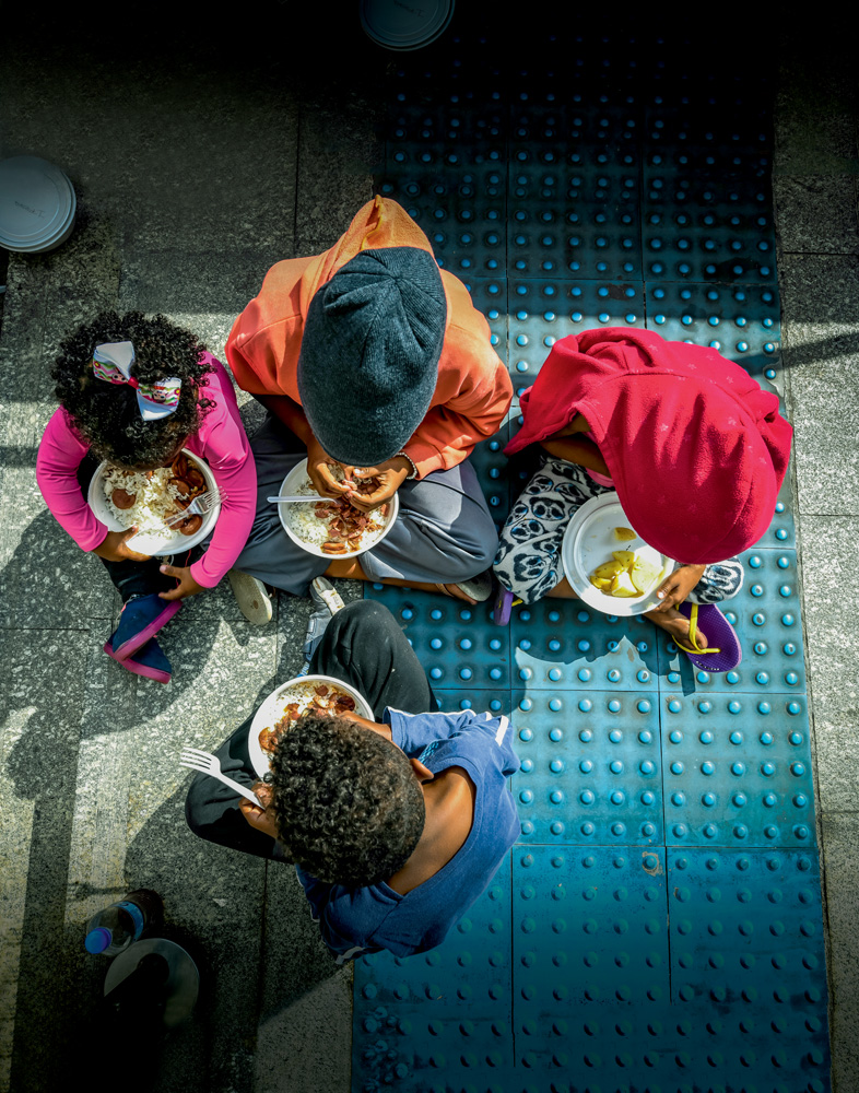 A imagem mostra quatro crianças almoçando em uma calçada em SP. A câmera tira a foto de cima, em que os rostos não aparecem, apenas a parte de cima da cabeça.