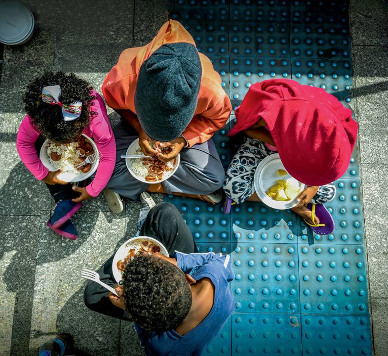 A imagem mostra quatro crianças almoçando em uma calçada em SP. A câmera tira a foto de cima, em que os rostos não aparecem, apenas a parte de cima da cabeça.