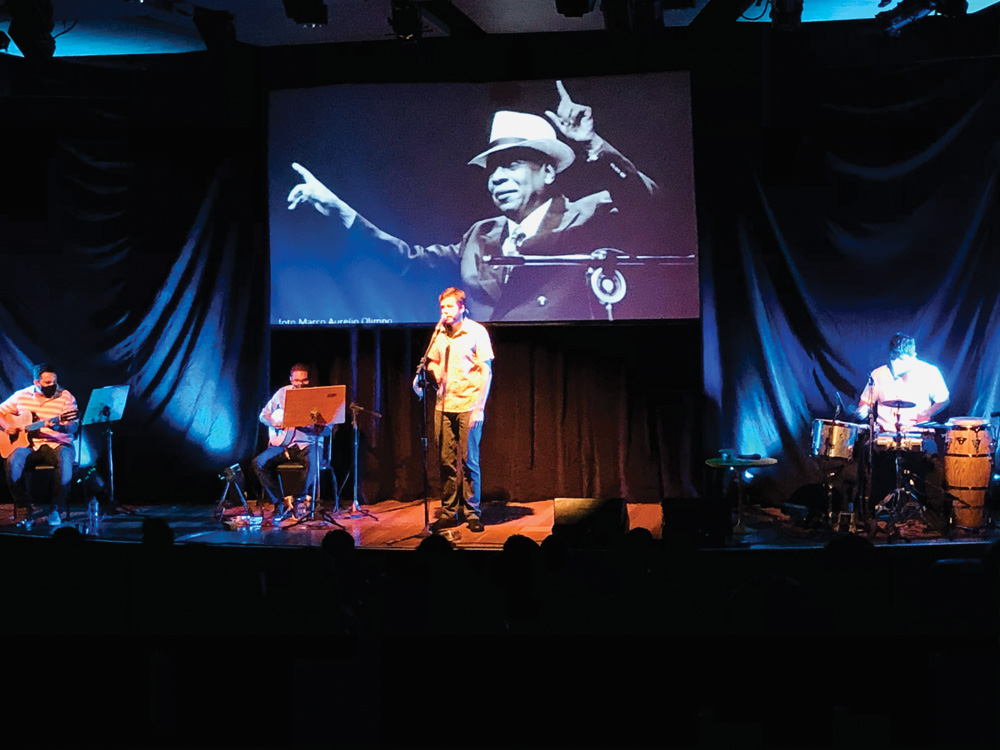 Em um palco, um homem canta com microfone, outros dois tocam violão e bateria e, ao fundo, um telão passa uma foto do cantor Zé Kéti