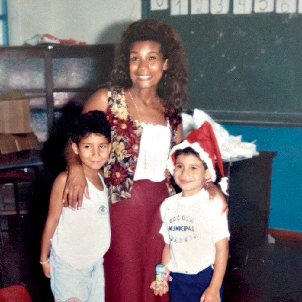 A imagem mostra a mãe de Konrad, Áurea Cunha, com mão nos ombros de duas crianças, com os três sorrindo para a câmera. Ao fundo, há uma lousa verde mostrando que eles estão em uma sala de aula.